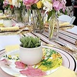 A table with floral plates, yellow napkins, a ceramic flower pot with drainage and bamboo tray, and bright flower centerpieces.