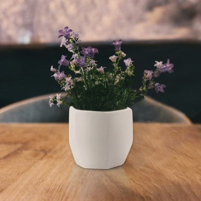 White Geometric Matte Pot with small purple and white flowers on a wooden table, modern decor setting with blurred chairs and soft lighting.